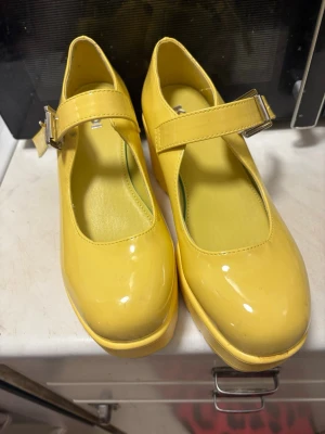 Gula Mary Jane ballerinaskor med klack - Säljer ett par glansiga gula Mary Jane må ballerinaskor med chunky klack och justerbart spänne i silver. Skorna har rund tå och plattformsula för extra höjd. Perfekta för dig som vill sticka ut med färg och retrovibe.
