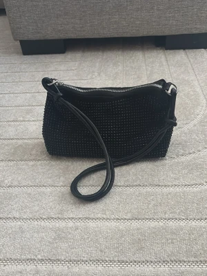 Svart axelväska med paljetter - Säljer en svart axelväska med glittriga paljetter och en smidig dragkedja upptill. Väskan har en enkel, rektangulär form och ett justerbart axelband. Perfekt för att addera lite extra stil till din outfit.