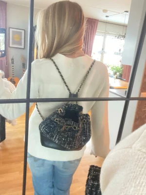 Svart och blå ryggsäck från Tiamo - Säljer en snygg ryggsäck från Tiamo med svart och blått rutigt tyg, detaljer i guld och svart skinn i botten. Väskan har kedjeaxelband och snörning upptill. Perfekt storlek för att få med allt viktigt och har en cool, trendig look.