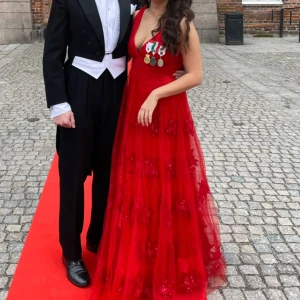 Röd långklänning med blommiga paljetter - Säljer en magisk röd långklänning med djup v-ringning och breda axelband. Klänningen har ett lager av transparent tyg dekorerat med blommiga paljetter som ger extra wow-effekt. Passformen är normal och materialet känns lätt och festligt, perfekt för bal!