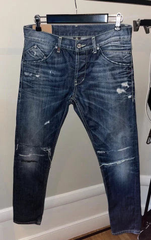 Blå distressed jeans från DSQUARED2 - Säljer ett par blå jeans från dondup med coola slitningar och trasiga detaljer på både fram- och baksida. Jeansen har raka ben, klassisk femficksdesign och kontrastsömmar. Tillverkade i Italien av högkvalitativt denim.