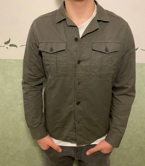 Grön overshirt från Riley, slim fit - Snygg militärgrön overshirt från Riley i slim fit-modell. Skjortan har två bröstfickor med knappar, klassisk krage och knäppning framtill. Tillverkad i mjuk bomull som är skön mot huden. Perfekt att bära över en t-shirt för en chill look.