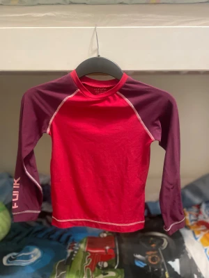Röd långärmad tröja från Funk - Snygg röd långarmad tröja/ underställ från Funk med kontrasterande mörklila armar och vita sömmar. Tröjan är gjord i mjuk polyester med lite stretch för skön passform. Perfekt för dig som gillar sportig stil och vill sticka ut med färg.  Barn till tjejer 130/140