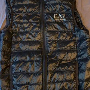 Svart dunväst från EA7 Emporio Armani - Snygg svart dunväst från EA7 Emporio Armani med quiltad design och glansig finish. Västen har dragkedja framtill och logga på bröstet. Perfekt att slänga över en hoodie för en chill look. Materialet är lätt och känns skönt mot kroppen och säljer p grund av att d blir vinter den har inte varit använd så mycket typ 10 gånger 