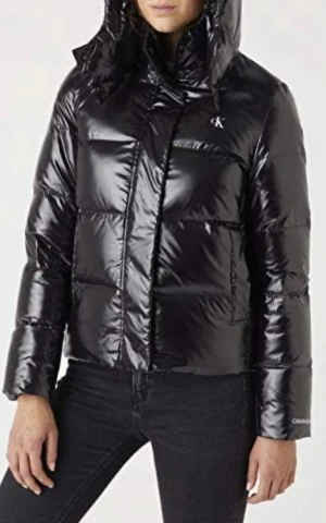 Svart jacka Calvin Klein Jeans - Svart glansig pufferjacka från Calvin Klein Jeans med huva och CK-logga på bröstet. Jackan har långa ärmar, dragkedja och knappar framtill. Använd en säsong, inga defekter. Lite mindre i storleken så passar XS-S