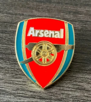 Arsenal pin i rött och guld - Cool Arsenal pin med klubbens klassiska logga i rött, guld och blått. Sköldformad design med tydlig text och kanonmotiv i guld. Perfekt för att visa att du är Arsenal supporter på jackan eller väskan.