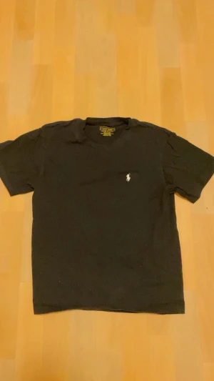 Svart t-shirt från Ralph Lauren - Svart t-shirt från Ralph Lauren med klassisk passform och korta ärmar. Ikoniska vita Polo-loggan broderad på bröstet. Tillverkad i mjuk bomull som känns skön mot huden. Perfekt för dig som gillar stilrena och enkla plagg med en touch av lyx.