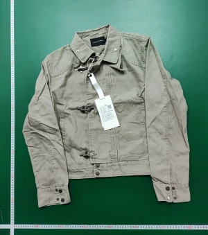 Beige overshirt med metallspännen - Snygg beige overshirt från Random Identities med coola metallspännen framtill och ståkrage. Jackan har långa ärmar med knappdetaljer vid mudden och är tillverkad i bomull. Perfekt för dig som gillar minimalistisk och modern stil.