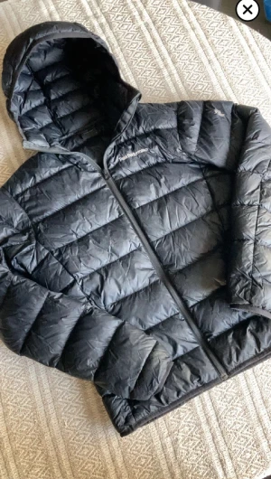 dunjacka Peak Performance  - Svart dunjacka från Peak Performance med huva och dragkedja framtill. Jackan har ett quiltat mönster och logotyp på bröstet. Perfekt för kalla dagar, snygg och stilren design som funkar till streetwear. Jackan är normal i passningen och har långärmade ärmar.