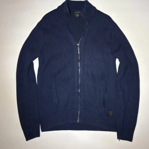 Marinblå stickad kofta från CKJ - Snygg marinblå stickad kofta från Calvin Klein Jeans med ribbad struktur och hel dragkedja framtill. Tröjan har två fickor och en liten CKJ-logga nedtill. Perfekt för dig som gillar stilrena och klassiska plagg med lite edge.