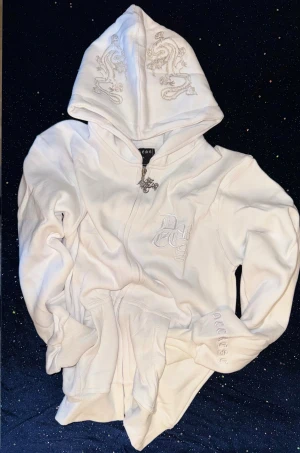 Vit Decrese Hoodie – Streetwear  XS - Decrese Sweden Hoodie i vitt. Exklusiv unisexmodell i mjuk bomull med lätt stretch och stentvättad finish. Vit broderad logga fram och silveraktiga detaljer på huva och rygg. Lite Oversize passform och två framfickor. Mått: axel 46cm, bröst 46 cm, längd 59cm, ärm 50 cm.‼️Köparen betalar retur och ny frakt vid byte‼️