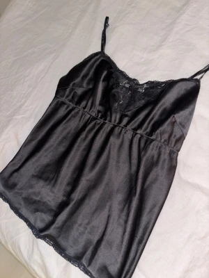 Svart satin linnetopp med spets - Snygg svart linnetopp i satin med tunna axelband och spetsdetaljer vid halsringningen och nederkanten. Toppen har en mjuk, glansig yta och en feminin v-ringad form. Köpt på Gina tricot🧸