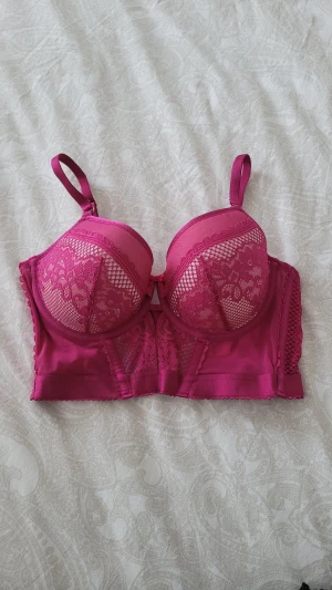 Rosa spetskorsett från Hunkemöller - Säljer en snygg rosa korsett från Hunkemöller i storlek 32DD/70E. Toppen har vadderade kupor, justerbara axelband och vackra spetsdetaljer framtill. Materialet är mjukt och stretchigt med mesh och spets för extra stilpoäng. Perfekt för dig som vill ha något extra.