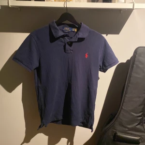 Marinblå pikétröja Polo Ralph Lauren - Snygg marinblå pikétröja från Polo Ralph Lauren i slim fit. Klassisk krage, korta ärmar och röd broderad logga på bröstet. Tillverkad i mjuk bomull som känns skön mot huden. Perfekt för dig som gillar stilrena och tidlösa plagg. Tröjan är nödgande helt oanvänd och i mycket bra skick.
