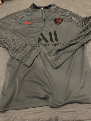 PSG långärmad träningsöverdel Nike - Snygg grå långärmad PSG-fotbollströja från Nike med halv dragkedja, svarta och röda detaljer samt diskret mönster på ärmarna. Tillverkad i lätt och ventilerande funktionsmaterial, perfekt för träning eller match.