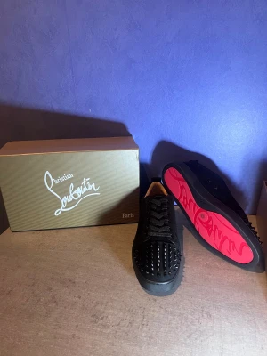 Christian Louboutin svarta sneakers med nitar - Svarta sneakers från Christian Louboutin med coola svarta nitar på tån och klassisk röd sula med logga. Skorna har snörning och är tillverkade i mocka med skinnfoder. Perfekt för dig som vill sticka ut med en edgy och lyxig stil.