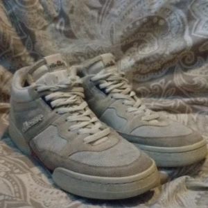 Grå sneakers från Ellesse - Snygga grå sneakers från Ellesse med högt skaft och klassisk retrostil. Skorna har snörning, rund tå och är tillverkade i en mix av mocka och textil. Perfekta för dig som gillar streetwear och vill ha en stilren look.