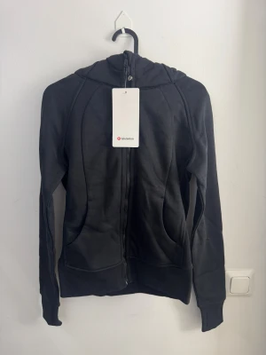 Svart slim fit hoodiejacka från Lululemon storlek 4 - Svart hoodiejacka från Lululemon med dragkedja framtill och huva. Tillverkad i mjuk bomulls- och polyestermix som känns skön mot kroppen. Snygg, figurnära passform och diskret logga på huvan. Perfekt för träning eller chill dagar.
