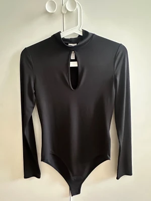 Svart långärmad bodysuit från H&M - Säljer en svart långärmad bodysuit med hög hals och snygg cut out-detalj framtill. Materialet är mjukt och stretchigt polyester med tight passform som sitter snyggt och ger en clean look. Perfekt att styla med jeans eller kjol