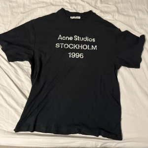 Svart Acne Studios t-shirt Stockholm - Svart t-shirt från Acne Studios med vit text 'Acne Studios STOCKHOLM 1996' på bröstet. Klassisk rund hals och korta ärmar. Materialet är mjuk bomull och passformen är normal. Perfekt för dig som gillar stilrena och ikoniska plagg. Passar dig som är runt 170-180cm ungefär