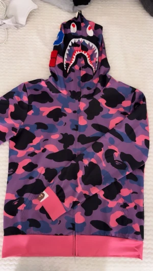 BAPE Shark Hoodie i lila camo - Säljer en BAPE Shark Hoodie med ikoniskt camouflagemönster i lila, rosa, blått och svart. Hoodien har dragkedja hela vägen, broderade bokstäver på huvan och detaljer som tiger och hajansikte. Muddar och nederkant i rosa, materialet är polyester. Perfekt statement-plagg!