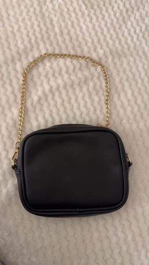 Svart axelväska med guldkedja - Snygg svart axelväska i skinnimitation med stilren, rektangulär form och guldfärgad kedja som axelrem. Väskan har dragkedja upptill och ett rymligt fack för dina essentials. Perfekt för dig som gillar minimalistisk och clean design.