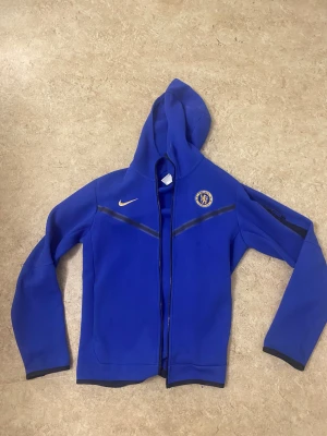 Chelsea FC blå vindjacka från Nike - Blå vindjacka med huva från Nike, prydd med Chelsea FC-logga och guldfärgad swoosh på bröstet. Jackan har hel dragkedja framtill, svarta detaljer och långärmad design. Perfekt för dig som älskar fotboll och vill ha en sportig look.