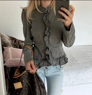 Kappa med volangdetaljer - Snygg kavaj med volang längs knäppningen och nederkanten. Figurnära modell med långa ärmar och markerad midja. Perfekt för dig som vill ha en unik twist på din outfit.