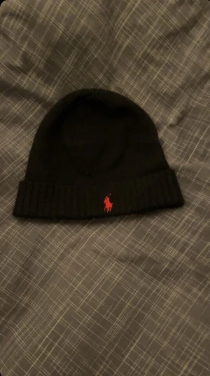 Svart mössa från Polo Ralph Lauren - Svart ribbstickad mössa från Polo Ralph Lauren med klassisk röd broderad logga framtill. Mjuk och varm, perfekt för kyliga dagar. Enkel design med uppvikt kant som ger en clean look.