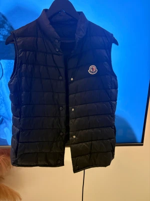 Mörkblå dunväst från Moncler - Svart quiltad dunväst från Moncler med klassisk logga på bröstet. Västen har ståkrage, tryckknappar framtill och horisontella sömmar som ger en snygg puffad look. Perfekt att slänga över en hoodie för extra värme och stil. Slitage finns då knappen längst ner har lossnat därav priset.