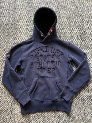 Mörkblå Superdry hoodie XS - Snygg mörkblå hoodie från Superdry i storlek XS med broderad text på bröstet och klassisk känguruficka. Tröjan har huva med mjukt material som känns skönt mot huden. Perfekt för dig som gillar sportig och avslappnad stil. Passar mig som är 174cm