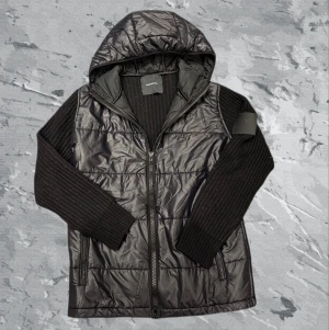 Svart hybridjacka Peak Performance M - Svart hybridjacka från Peak Performance med quiltad, glansig framsida och huva i syntetmaterial samt stickade, ribbade ärmar och rygg. Jackan har dragkedja framtill och en cool, sportig look som funkar till streetwear. Perfekt för dig som gillar stil och funktion.