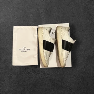 Valentino Garavani Open Sneakers 43 - Säljer ett par Valentino Garavani Open sneakers i vitt med bred svart läderdetalj över mitten. Skorna har rund tå, platt sula och snörning. Tillverkade i kalvskinn och kommer med original dustbag och låda. Perfekt för dig som gillar lyxiga och stilrena sneakers.