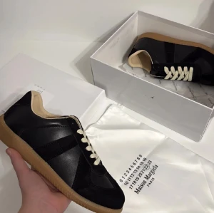 Maison Margiela Replica svarta sneakers - Maison Margiela Replica sneakers i svart skinn och mocka med beige detaljer och snörning. Klassisk rund tå och platt gummisula i ljusbrun färg. Snygg minimalistisk design med kontrasterande vita skosnören och diskret branding på sulan.
