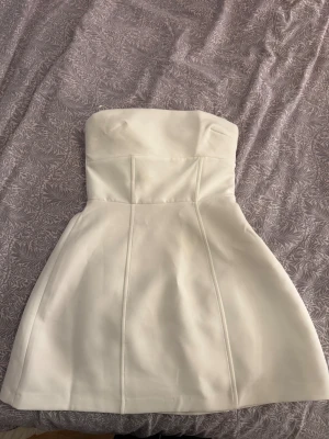 Vit axelbandslös kort klänning med rosett - Supersöt vit axelbandslös miniklänning med  och en stor rosett baktill. Klänningen har en lätt utsvängd kjol och är perfekt för dig som vill sticka ut med en söt och stilren klänning. Aldrig använd💓inköp för 460kr.