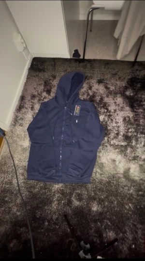 Marinblå hoodie från Polo Ralph Lauren - Snygg marinblå hoodie från Polo Ralph Lauren med dragkedja framtill och klassisk huva med snörning. Tröjan har lång ärm och den ikoniska lilla polospelaren broderad på bröstet. Perfekt för chill dagar och streetwear-stil.
