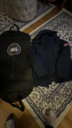 Mörkblå Canada Goose dunjacka - Säljer en mörkblå dunjacka från Canada Goose med klassisk patch på ärmen. Jackan har lång ärm, huva och stängs med både dragkedja och knappar. Perfekt för kalla vinterdagar och levereras med original fodral. Riktigt snygg och tidlös modell. BOX MED