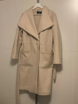 Beige kappa med bälte från Bik Bok - Säljer en stilren beige kappa från Bik Bok med bred krage och två stora fickor framtill. Jackan har ett bälte i midjan för snygg passform! 