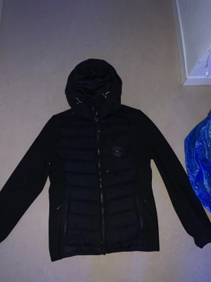 Svart pufferjacka från Zavetti Canada - Svart pufferjacka från Zavetti Canada med huva och snygga quiltade paneler framtill. Jackan har dragkedja, två sidofickor med zip och broderad logga på bröstet. Materialmix med syntet och polyester för extra komfort och stil.