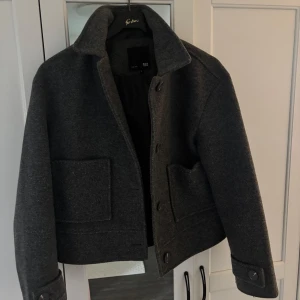 Mörkgrå ulljacka från 157 - Säljer en mörkgrå, kortare ulljacka från 157 med stora fickor framtill och knappar vid ärmslut. Jackan har klassisk krage och är helfodrad, perfekt för kyligare dagar. Den är använd ungefär i 2 månader men inte något fel alls på den. Jag är 178 cm lång och den sitter bra på mig:)