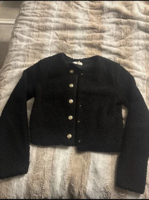 Svart teddyjacka med knappar - Säljer en svart teddyjacka med rund hals och snygga guldfärgade knappar framtill. Jackan är croppad och har lång ärm, perfekt för dig som gillar en chill och trendig look. Materialet är mjukt och fluffigt, vilket gör den extra mysig under höst och vinter.