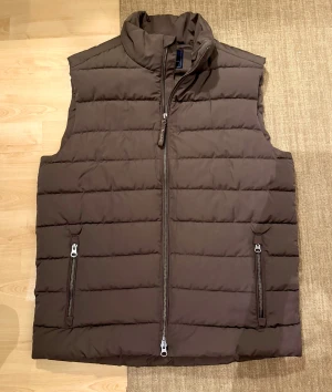 Brun dunväst från Bondelid, herr-modell strl S/M - Snygg brun dunväst från Bondelid med quiltad design och hög krage. Västen har två praktiska fickor med dragkedja och en stilren look. Perfekt att slänga över en hoodie eller tröja när det är kyligt ute. Köpt på MQ för 1199kr, helt ny och oanvänd. Storlek S herr-modell, passar till både S och M! 