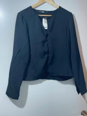 Svart blus med v-ringning från Bik Bok - Säljer en stilren svart blus från Bik Bok med v-ringad hals och långa ärmar. Blusen har en enkel, loose passform och är tillverkad i ett mjukt polyestermaterial. Perfekt för dig som gillar minimalistisk och clean stil.