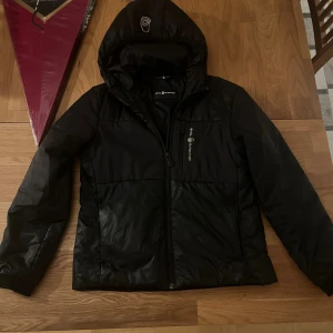 Svart pufferjacka från Sail Racing - Svart pufferjacka från Sail Racing med huva och dragkedja framtill. Jackan har logotyp på bröstet och huvan, samt två sidofickor med dragkedja. Materialet är syntet och jackan är långärmad med normal passform. Perfekt för kalla dagar. 