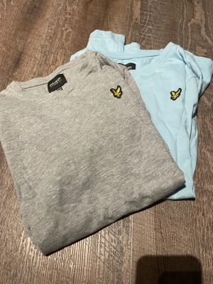 2-pack Lyle & Scott t-shirts junior - Två stilrena t-shirts från Lyle & Scott Junior, en i ljusgrått och en i ljusblått. Båda har korta ärmar och klassisk rund hals. På bröstet sitter den ikoniska gula örn-loggan. Materialet är mjuk bomull som känns skönt mot huden.