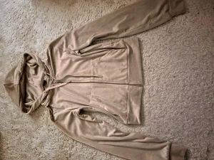 Beige croppad hoodie i velour - Säljer en beige croppad hoodie från Juicy Couture i mjuk velour. Tröjan har huva med snörning, dragkedja framtill och långa ärmar. Perfekt för en chill och trendig look. Snygg att matcha med jeans eller mjukisbyxor. 