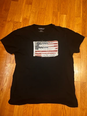 Svart t-shirt Ralph Lauren flagga M - Svart t-shirt från Ralph Lauren Denim & Supply i 100% bomull. Framsidan har en stor patch med amerikansk flagga och slitna detaljer samt texten 'Denim & Supply Ralph Lauren'. Klassisk passform och korta ärmar. Pris kan diskuteras!  Skriv vid frågor eller funderingar