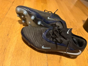Nike phantom 6 Elite ag  - Säljer ett par svarta Nike fotbollsskor med blå detaljer och ljusblå Swoosh. Skorna har snörning och ribbad ovandel i syntetmaterial för extra grepp. Sulan har fasta dobbar i ljusblått, perfekt för spel på gräs. Snygg, modern design med rund tå och låg profil.