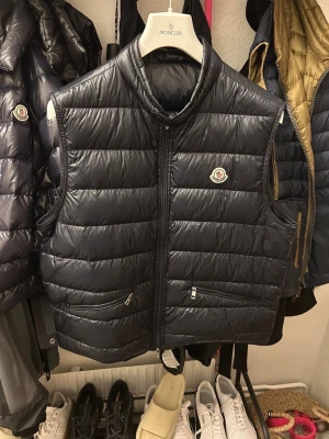 Svart dunväst från Moncler - Svart/navy dunväst från Moncler med klassisk quiltad design och hög krage. Västen har dragkedja framtill, två fickor med dragkedja och Moncler-logga på bröstet. Tillverkad i glansig polyester och dunfylld för extra värme. Perfekt för lager-på-lager-stil.