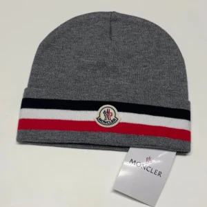 Grå Moncler mössa med ränder - Snygg grå mössa från Moncler med breda ränder i svart, vitt och rött runt kanten. Framtill sitter Monclers klassiska logotyp. Mössan är ribbstickad och har en clean, sportig look som passar perfekt till streetwear. Materialet är mjuk bomull.